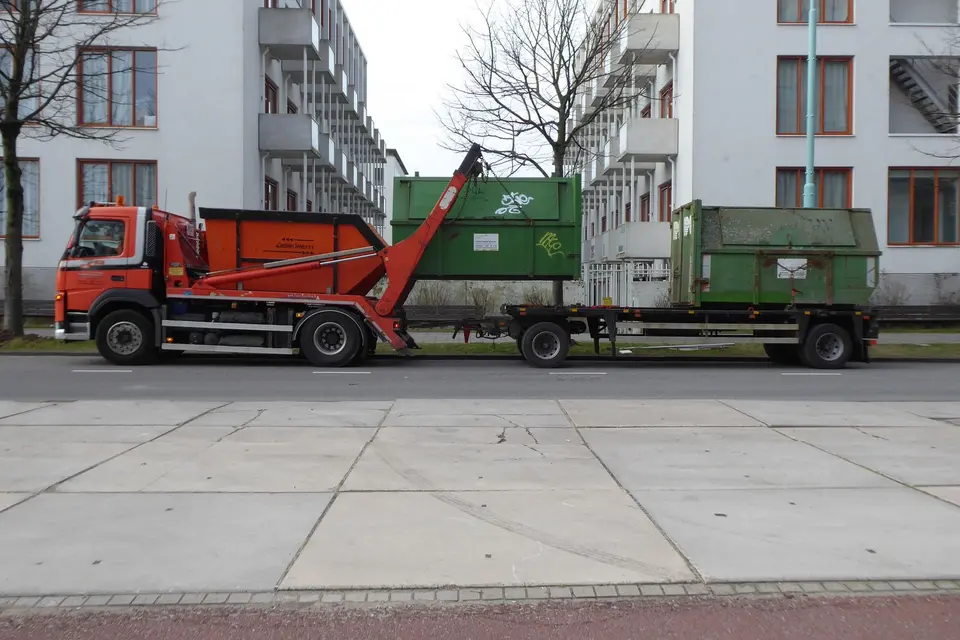 Camion porte-benne et remorque chargés de bennes à déchets, garés le long d'un trottoir